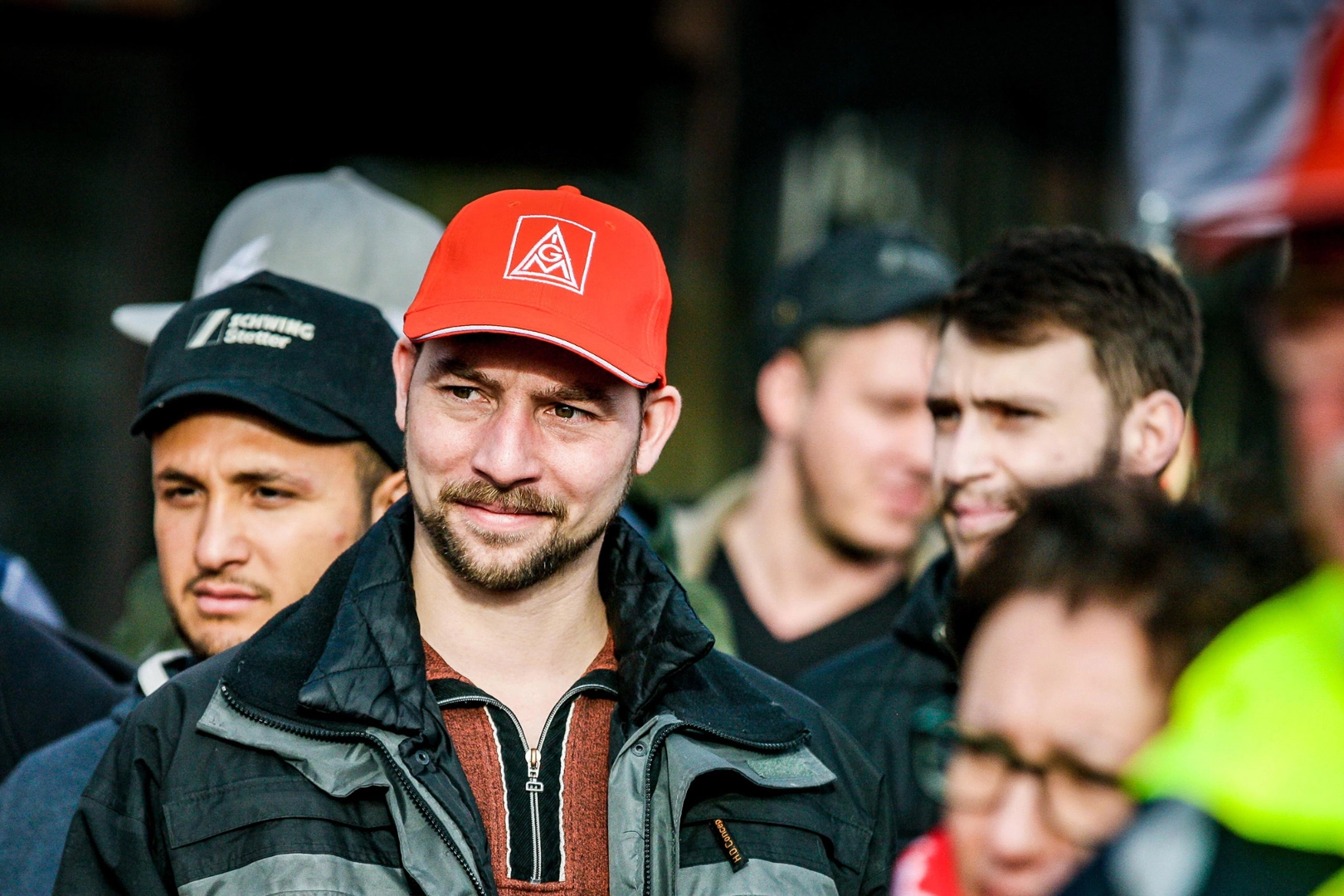 Ein junger Mann mit IG Metall-Kappe steht in eine Gruppe Menschen und lächelt.