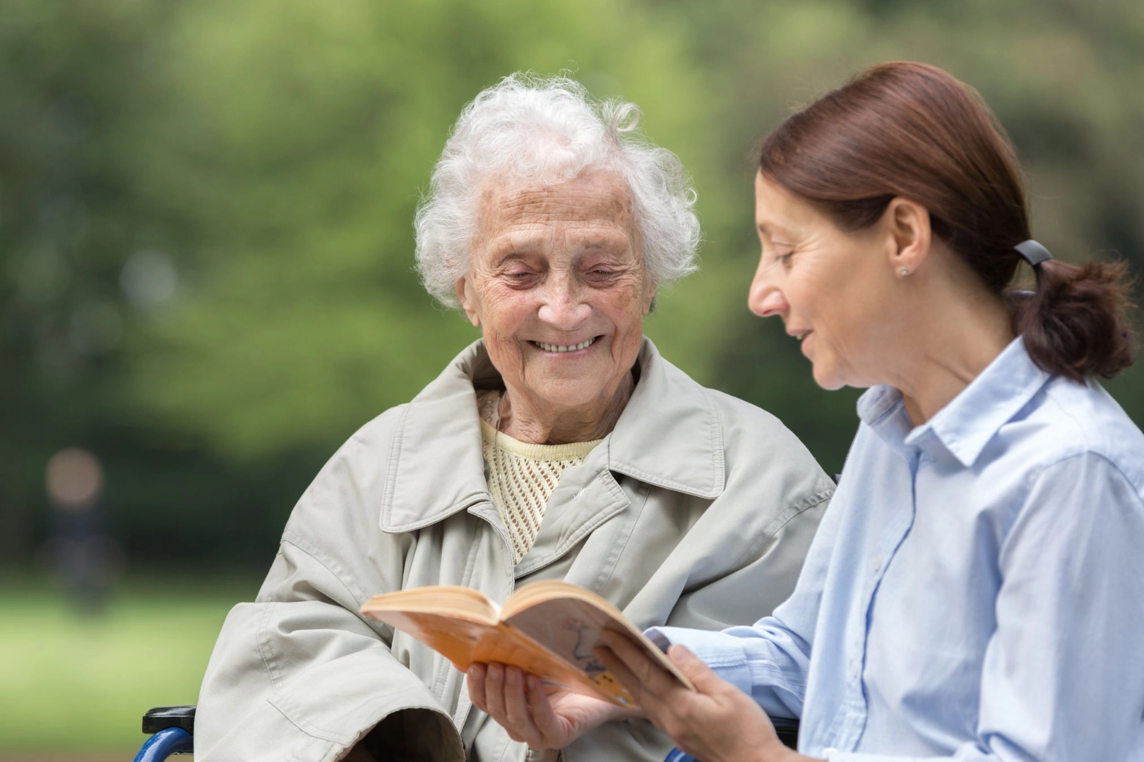 Eine Seniorin im Rollstuhl und ihre Tochter schauen in einem Park gemeinsam in ein Buch.