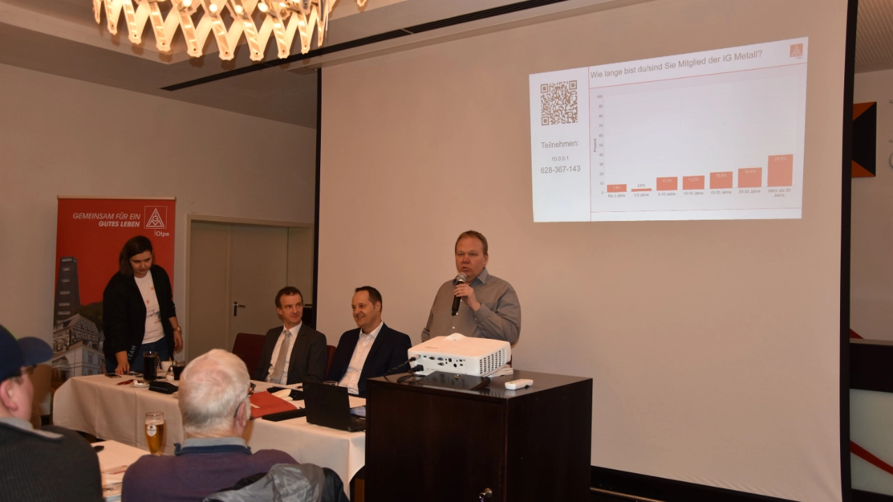 Andre Arenz begrüßt Wahlversammlung im Kolpinghaus Olpe zur ersten Wahlversammlung. Hinter ihm eine Leinwand mit Präsentation zum Thema.