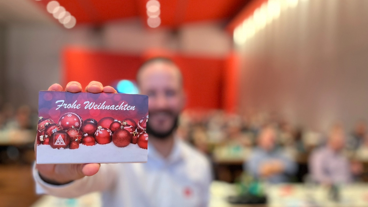 Wir sehen um Hintergrund einen verschwommenen Saal voller betrieblicher Gremien und einen IG Metall Sekretär im Vordergrund, der eine IG Metall Karte mit dem Aufdruck Frohe Weihnachten in die Kamera hält.