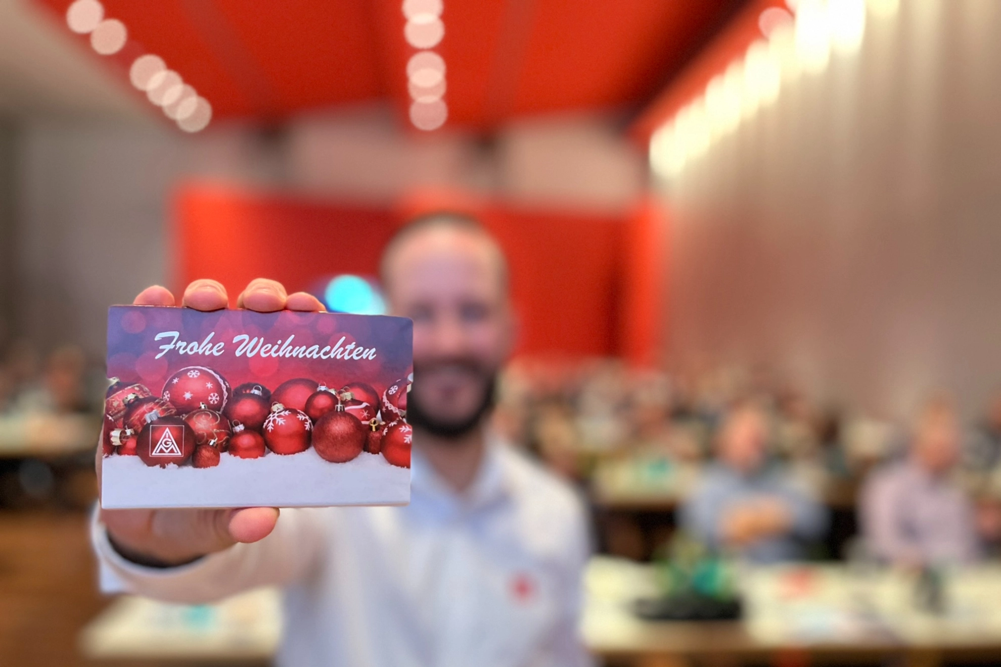 Wir sehen um Hintergrund einen verschwommenen Saal voller betrieblicher Gremien und einen IG Metall Sekretär im Vordergrund, der eine IG Metall Karte mit dem Aufdruck Frohe Weihnachten in die Kamera hält.