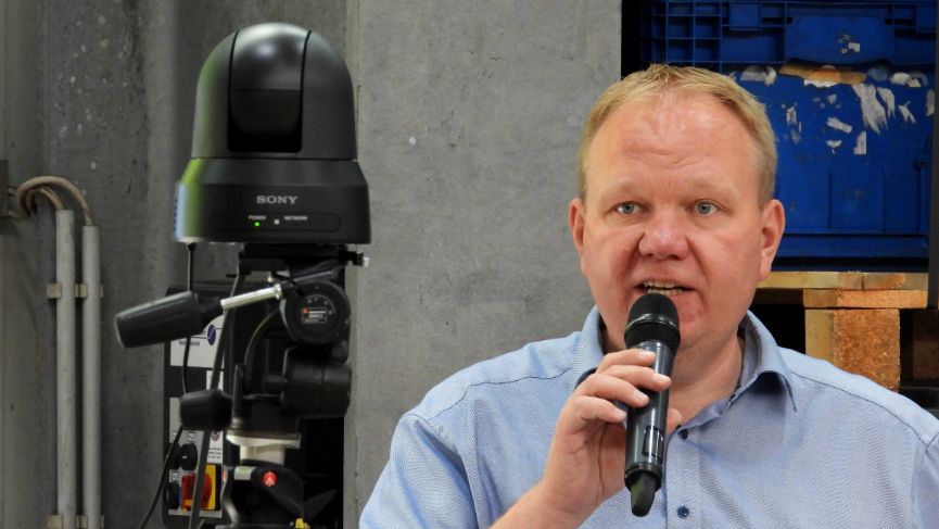 Andre Arenz spricht auf Betriebsversammlung bei KDK in eine Kamera und ist so im Saal und bei Teilnehmenden Zuhause sicht- und hörbar.