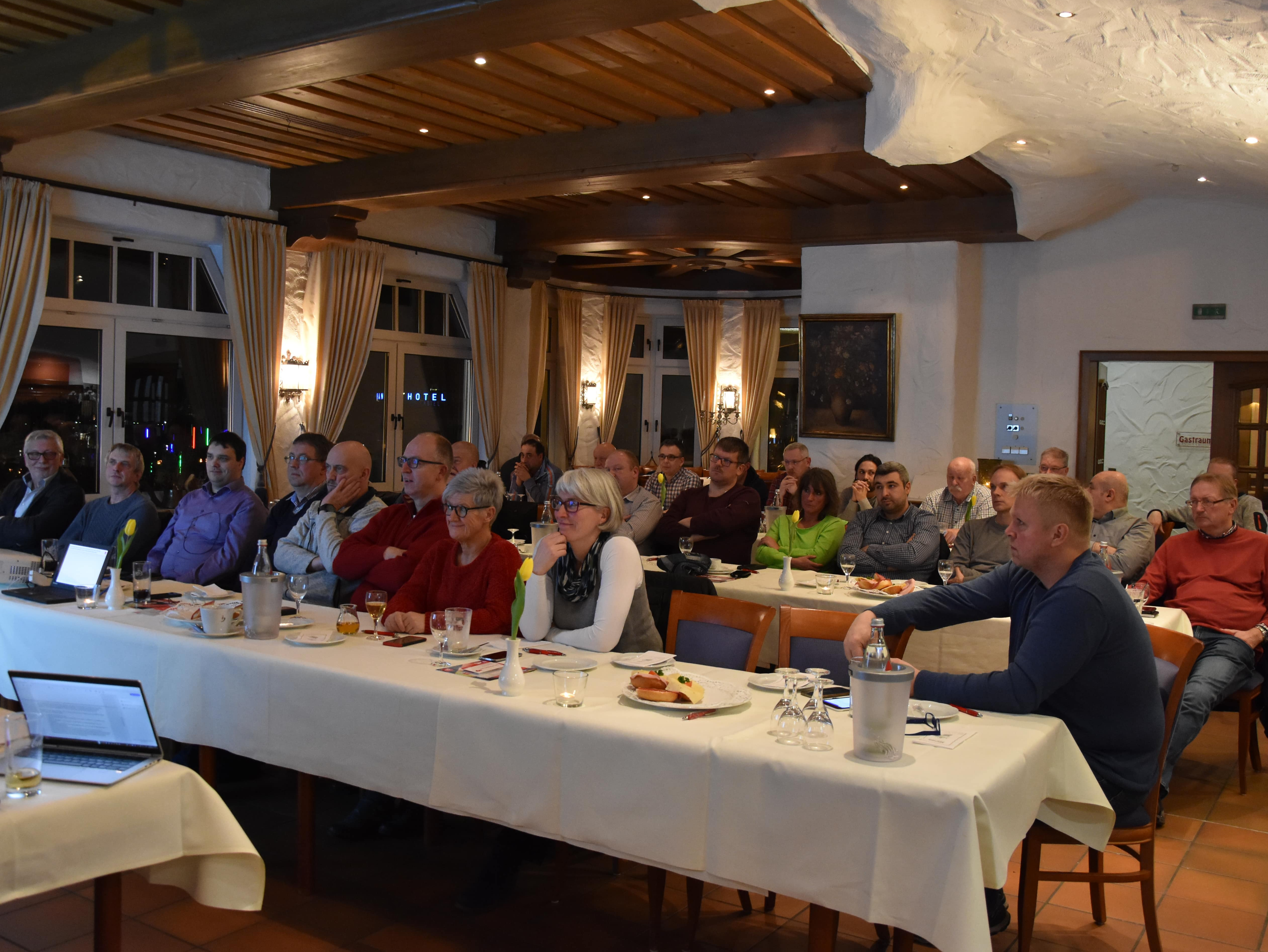 Kolleg*innen der Wahlversammlung schauen auf Leinwand und hören den Ausführungen von André Arenz zu.