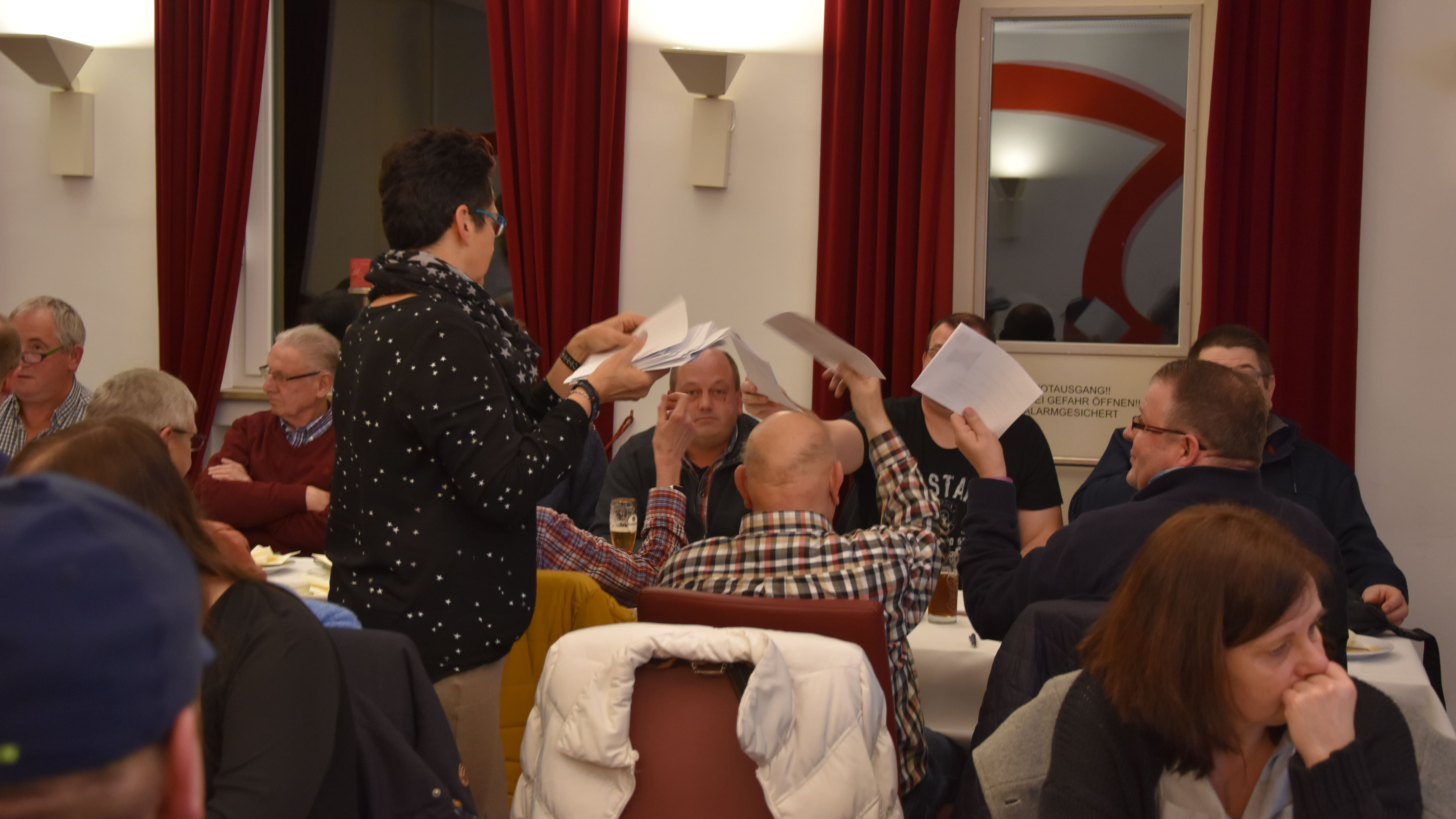 Eine Kollegin sammelt Stimmzettel von einer Gruppe Teilnehmender ein. Eine große Gruppe Menschen ist zu sehen, die ihr Wahlzettel entgegenstrecken.